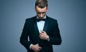 Elegancki mężczyzna w galowym stroju męskim, z czarną muszką i zegarkiem na ręku, poprawia marynarkę.
