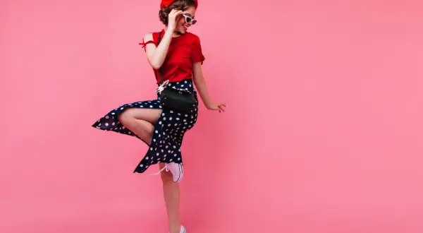 Dziewczyna w stylu pin-up girl w czerwonej bluzce, spódnicy w grochy i czerwonym beretce, zgrabnie podskakuje na różowym tle.