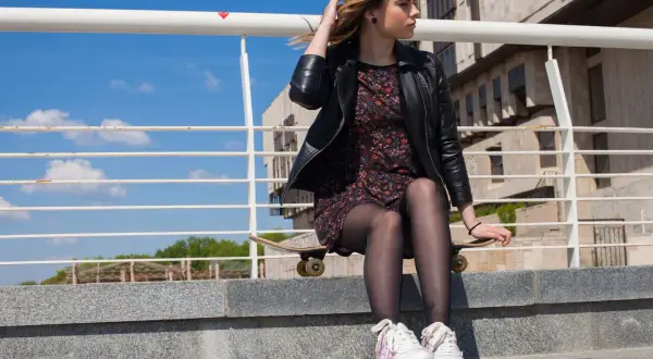 Dziewczyna w eleganckiej sukience i sportowych butach siedzi na deskorolce na schodach.