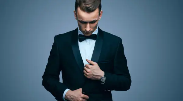 Elegancki mężczyzna w galowym stroju męskim, z czarną muszką i zegarkiem na ręku, poprawia marynarkę.