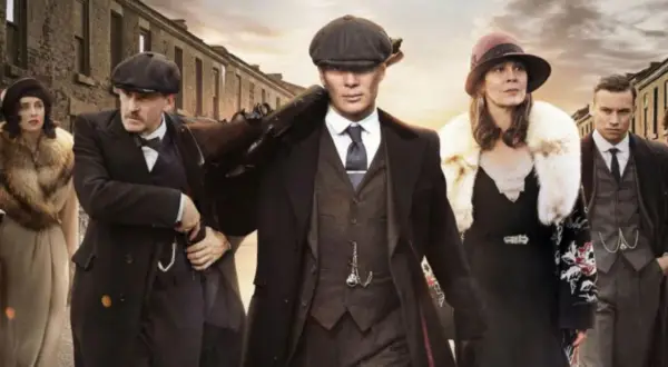 Gang w stylu Peaky Blinders, w tym Tommy Shelby z karabinem, idzie ulicą. Eleganckie płaszcze i kapelusze.