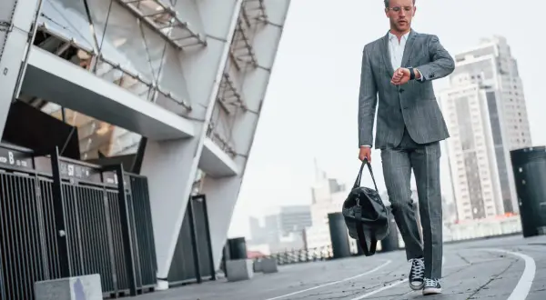Elegancki garnitur i trampki na wesele – młody mężczyzna sprawdza zegarek, idąc z torbą obok nowoczesnego budynku.