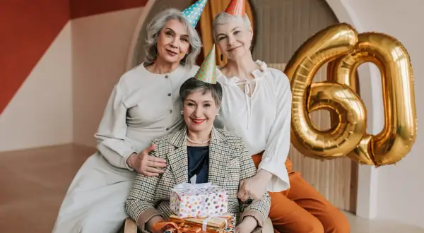 Eleganckie stylizacje na urodziny w lokalu. Trzy kobiety świętują 60. urodziny, otoczone prezentami i złotymi balonami.