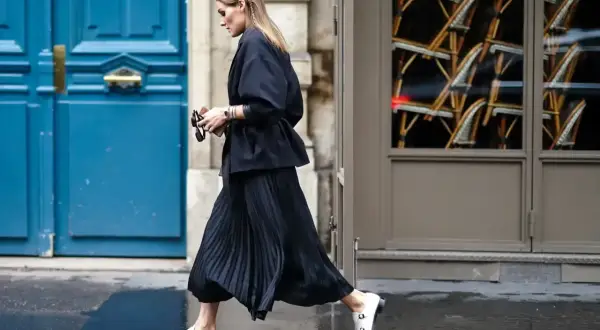 Elegancka kobieta w czarnej, plisowanej spódnicy i marynarce, tworząca stylową stylizację. Białe mokasyny dodają kontrastu.