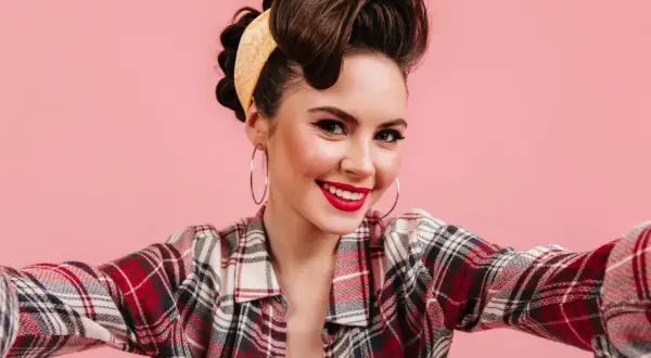 Uśmiechnięta pin-up girl w kratę i żółtej bandanie, z burzą loków i dużymi kolczykami, pozuje do zdjęcia.