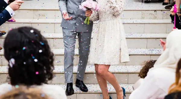 Eleganckie ślubne stylizacje pary młodej na schodach, obsypanej konfetti. Panna młoda w koronkowej sukience, pan młody w szarym garniturze.