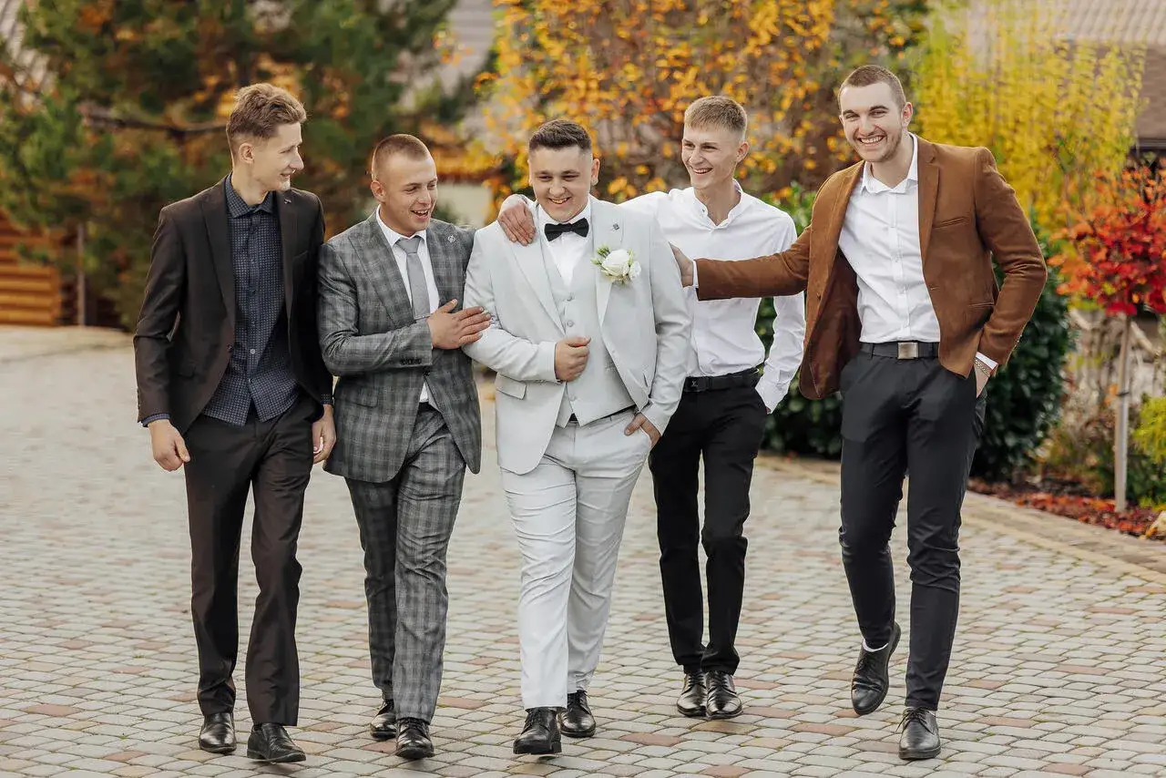 Pięciu mężczyzn w galowych strojach, w tym pan młody w jasnej marynarce, idzie razem, ciesząc się chwilą.