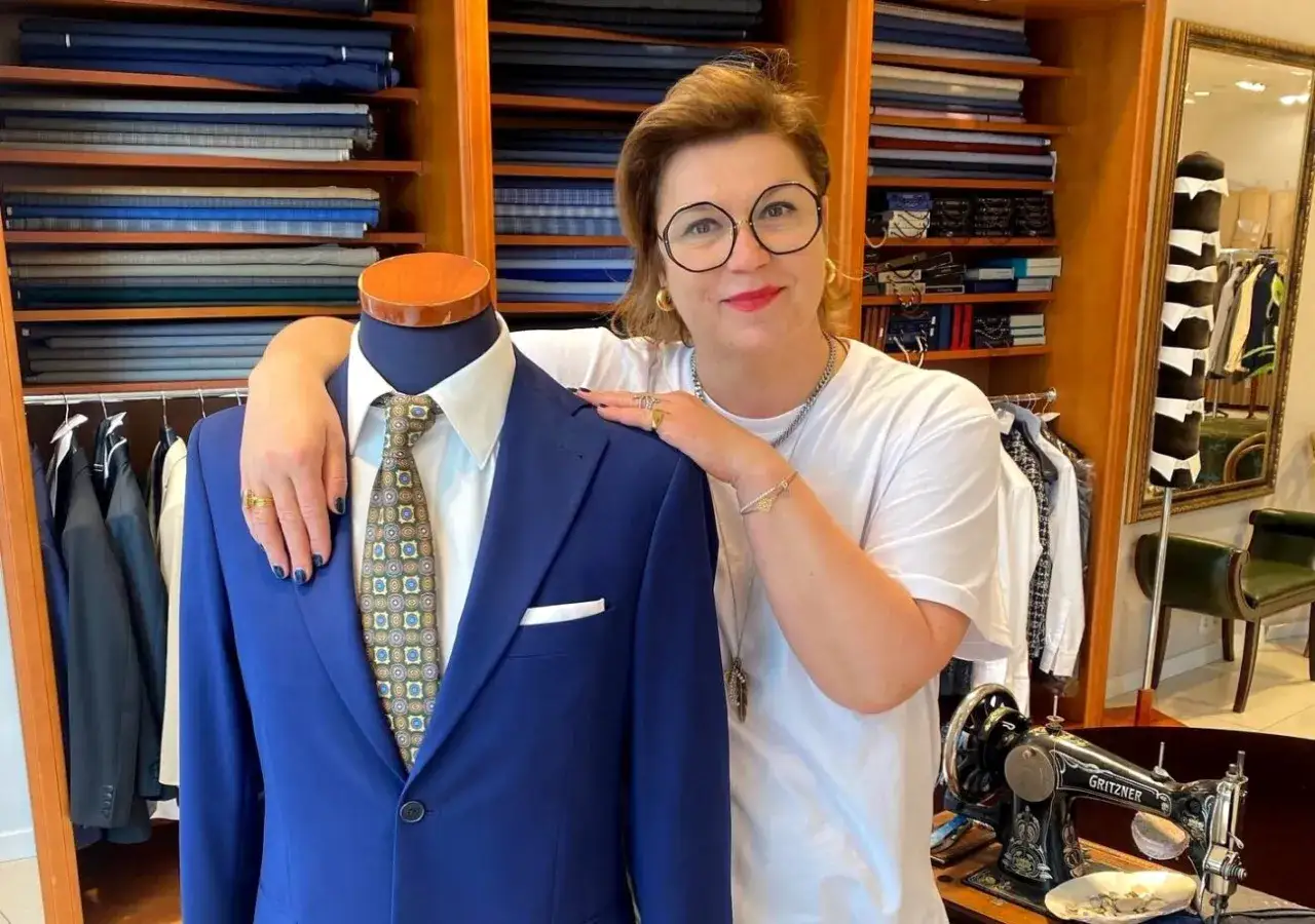 Kobieta w okularach, prawdziwy trend setter, prezentuje elegancki, niebieski garnitur na manekinie. W tle półki z tkaninami i stara maszyna do szycia.